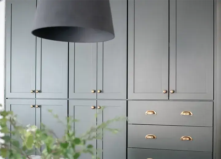 Green cabinet doors are seen in a wall of cabinets with brass knobs and cup pulls, and a large black pendant light.