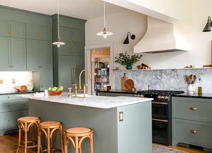 Kitchen Cabinet Doors A kitchen with muted green shaker style kitchen cabinet doors, wooden bar stools, marble countertops, a plaster range hood, brass hardware and dainty pendant lights.