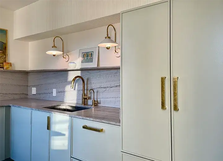 A picture of a contemporary apartment kitchen with trendy Skinny shaker cabinet doors in a soft-grey colour. Gold cabinet hardware, grey marble countertops are seen with two pendant lights flanking the sink.