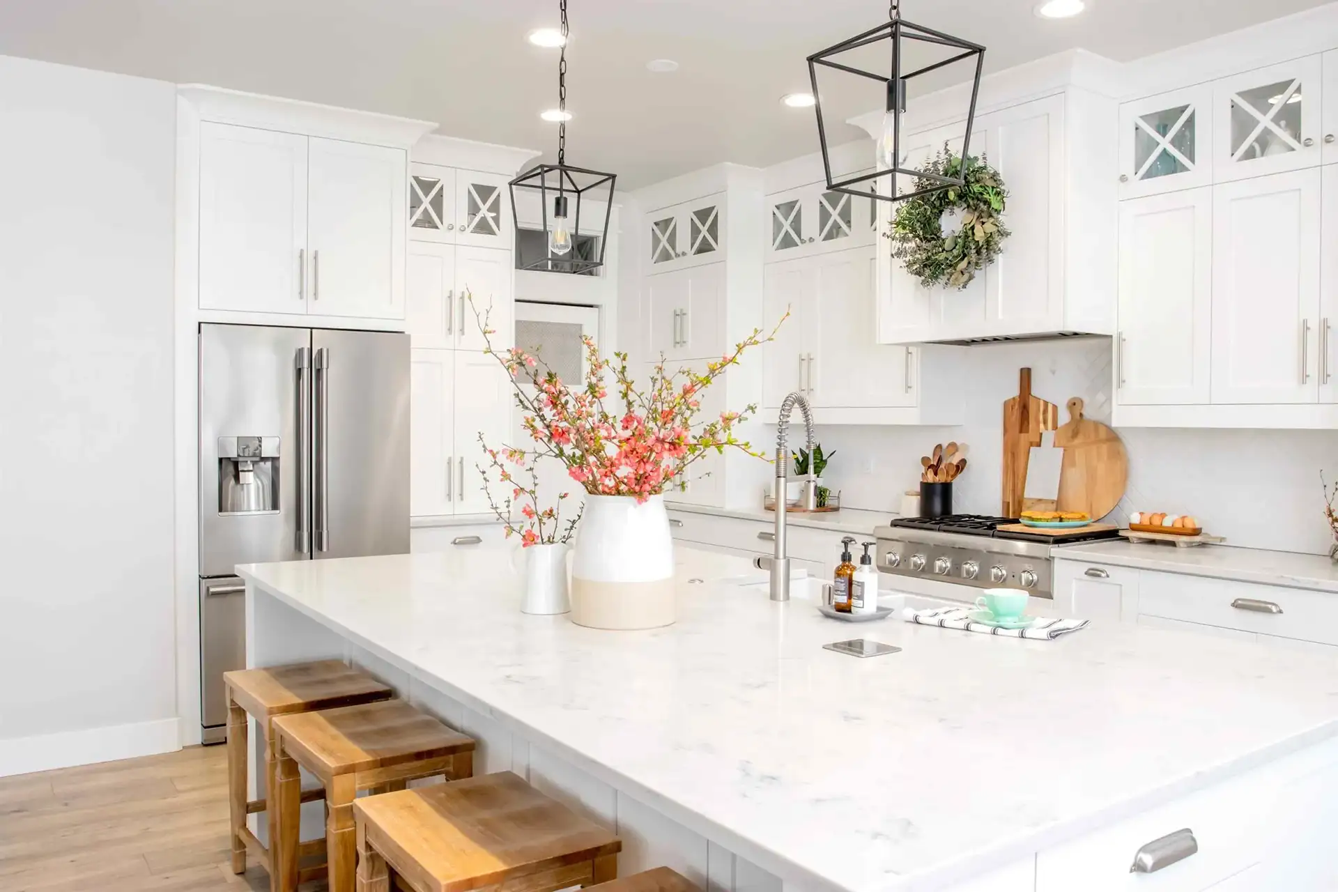 An image of a white kitchen with shaker cabinet doors, wooden bar stools, black lighting, nickel hardware, with decorative cutting boards and greenery on the countertop.