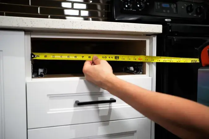 A photo of hands holding a yellow measuring tape up to a kitchen drawer opening.