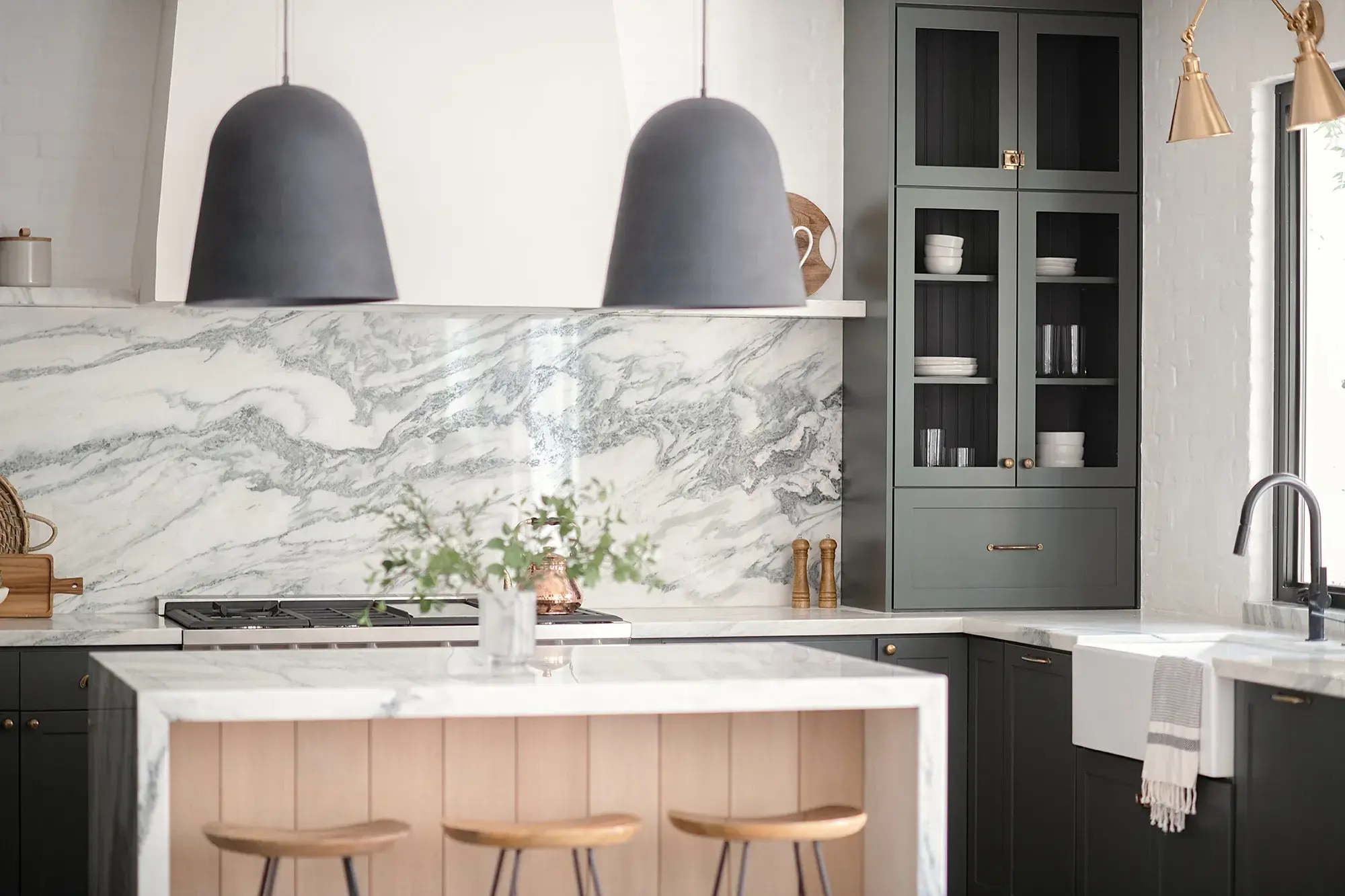 A photo of a kitchen with dark green cabinet doors, large black pendant lights, a plaster range hood, wooden island an waterfall countertop.