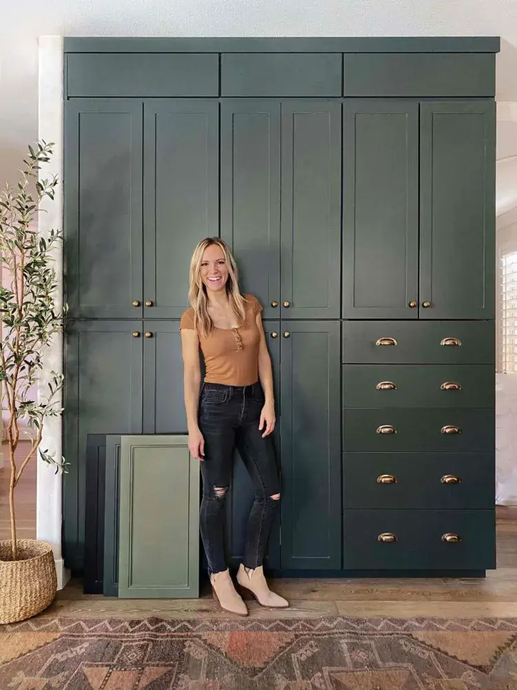 An image of Angela Rose standing in front of a wall of pantry cabinets in the Slim Shaker door in Dream Dusk, with doors resting on the ground.