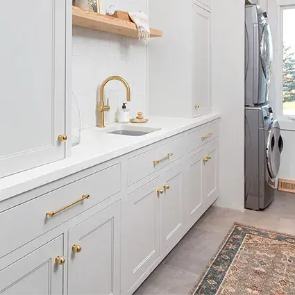 An image of a laundy room with beige stepped shaker shaker cabinet doors, faux concrete countertops, brass hardware, and wooden floating shelves.
