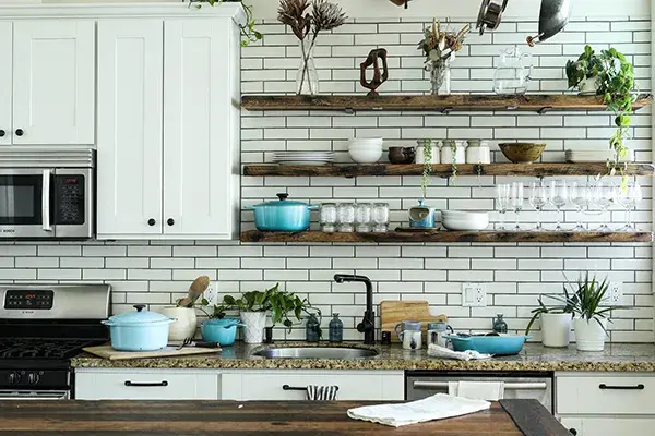 An image of a kitchen with white shaker cabinet doors, a white backsplash, and wooden floating shelves with black hardware.