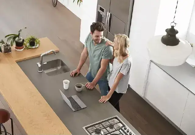 A photo of a man and woman standing at their kitchen counter with a laptop open, coffee mugs.