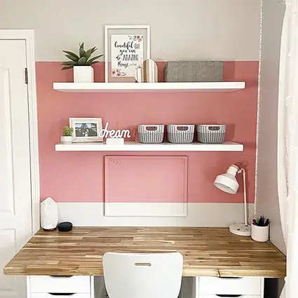 An image of two white floating shelves over a desk with wooden worktop, with a pink and white painted wall.
