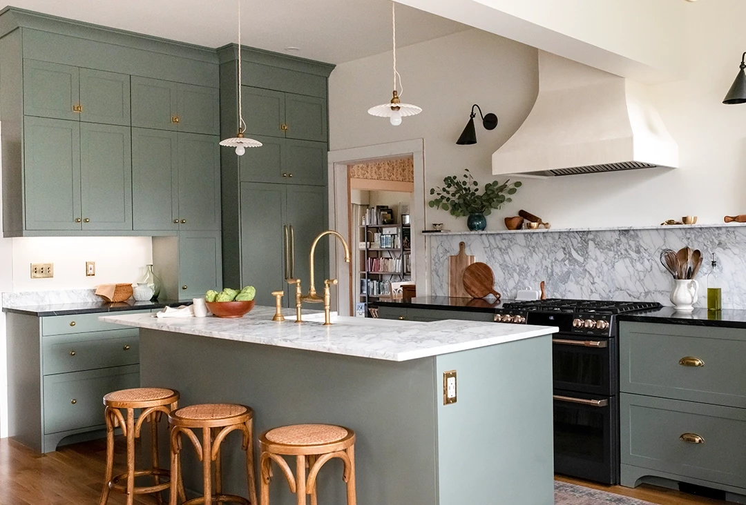 An updated kitchen with slim shaker cabinet doors in a soft green color, with marble countertops, a black range, plaster hood, and brass hardware.