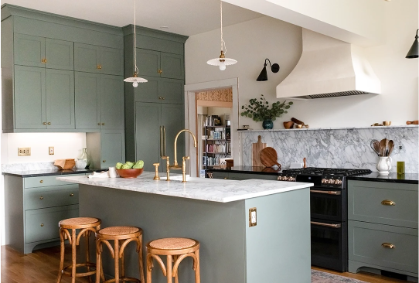 An updated kitchen with slim shaker cabinet doors in a soft green color, with marble countertops, a black range, plaster hood, and brass hardware.