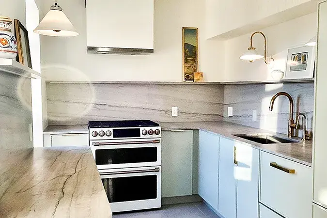 An image of a kitchen with skinny shaker cabinet doors in light grey with gold hardware. Marble countertops and a white plaster range hood.