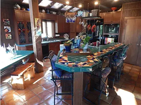 An image of an outdated kitchen in a lake house with tile countertops, oak cabinetry, and an Aztech inspired design.
