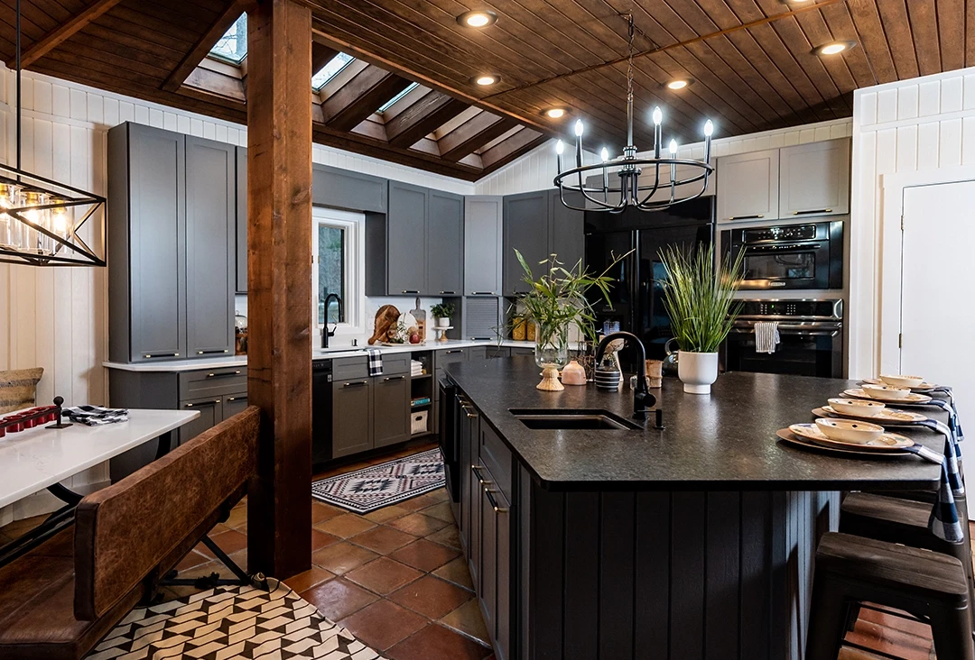 An image of a lakehouse kitchen with grey cabinet doors, wood ceiling and posts, quartz countertops, and black hardware.