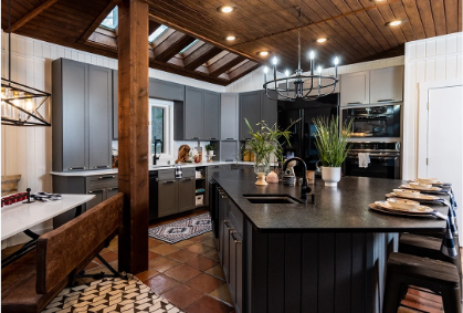 An image of a lakehouse kitchen with grey cabinet doors, wood ceiling and posts, quartz countertops, and black hardware.