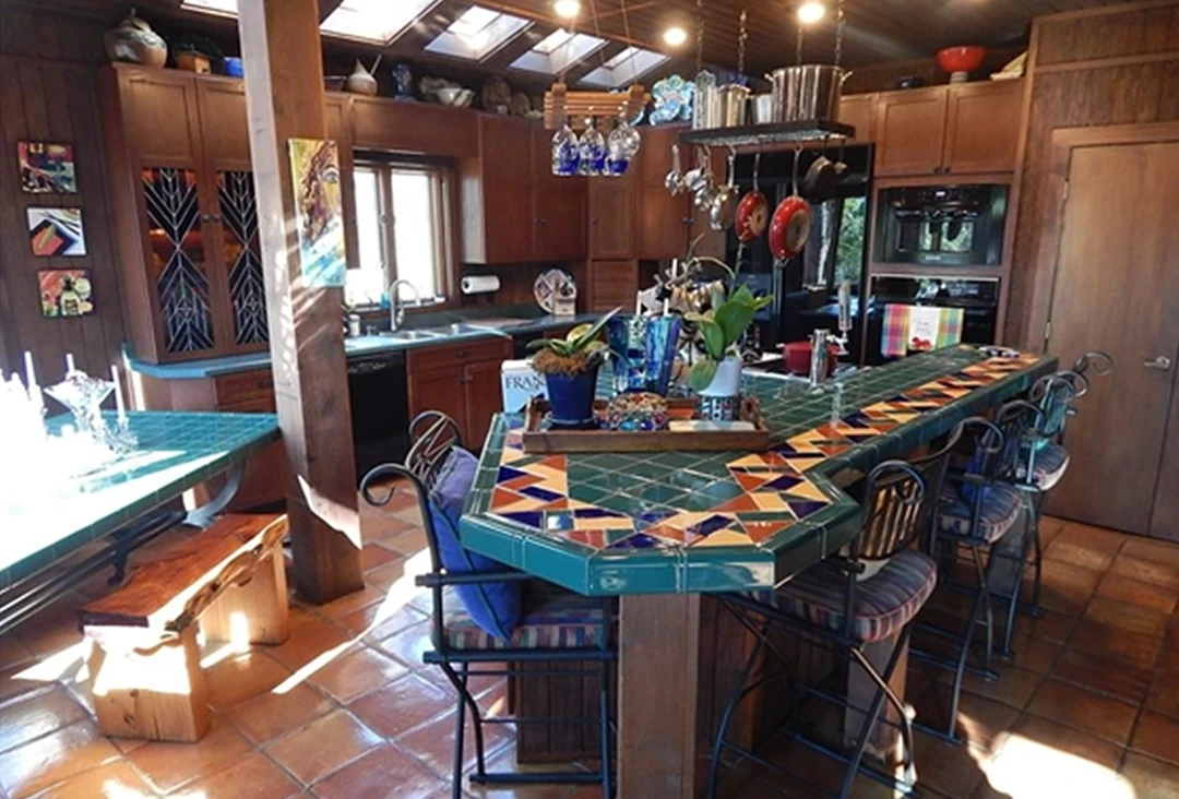 An image of a dated kitchen with honey oak cabinet doors, tile countertops, and a hanging pot rack.