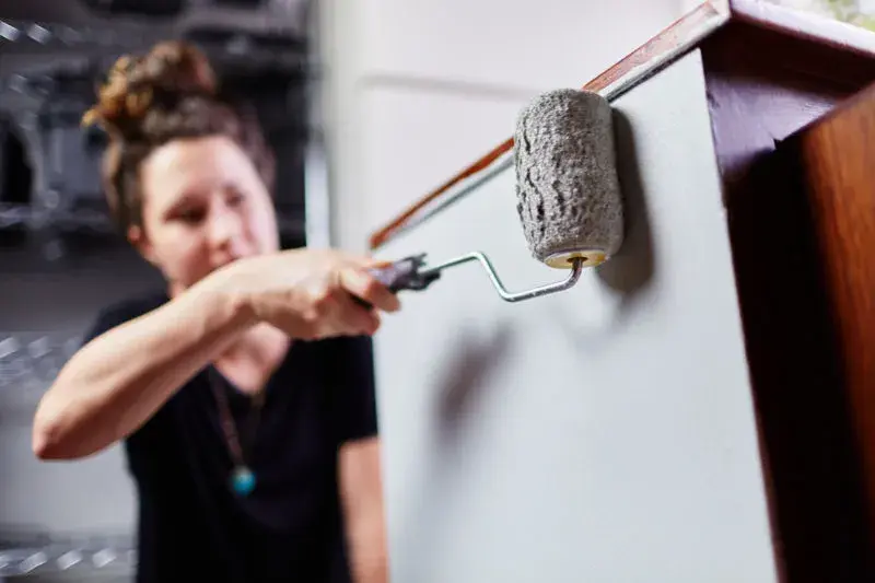 An image of a woman painting the side of a cabinet with a roller.
