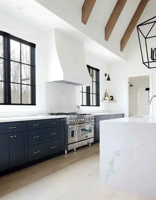 An image of a kitchen with vaulted ceilings, wooden beams, navy blue base cabinets, black windows, and a plaster range hood.
