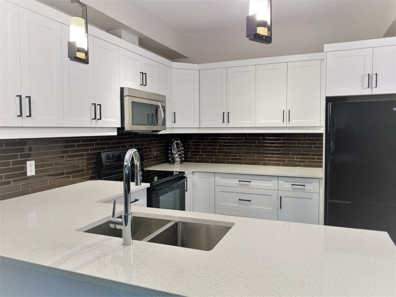 An image of a kitchen with white stepped shaker cabinet doors and speckled quartz countertop, black appliances, black hardware.