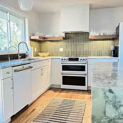 An image of a kitchen with white shaker cabinet doors, blue marble countertops, white appliances, gold hardware, and a plaster range hood with wooden floating shelves.