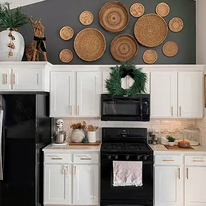 An image of a kitchen with white shaker cabinet doors, marble countertops, black appliances, and gold hardware. A wreath sits on two cabinet doors and a dark grey wall is covered with decorative baskets.