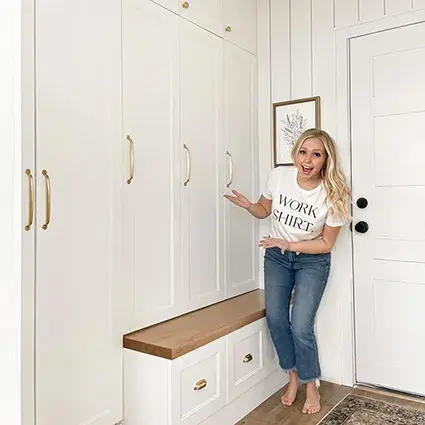 An image of a woman standing in front of white stepped shaker cabinet doors in a mudroom with wooden bench top and brass hardware.
