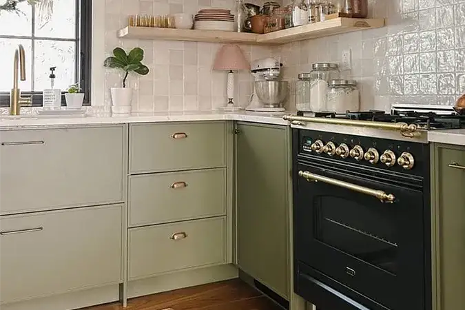 An image of a kitchen with olive green skinny shaker cabinet doors, a black and gold range, brass hardware, wooden floating shelves and a soft grey backsplash.