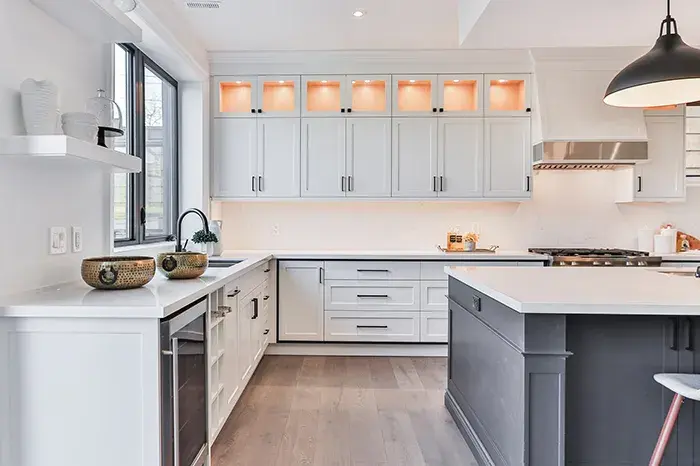 An image of a kitchen with white shaker style perimeter cabinets, glass upper doors, and a dark grey island. Black hardware is seen throughout the kitchen.