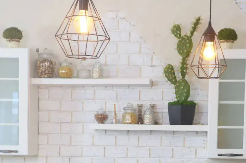 An image of a kitchen with white floating shelves, white brick on the wall, white upper cabinets, and decorative pendant lights and cacti.