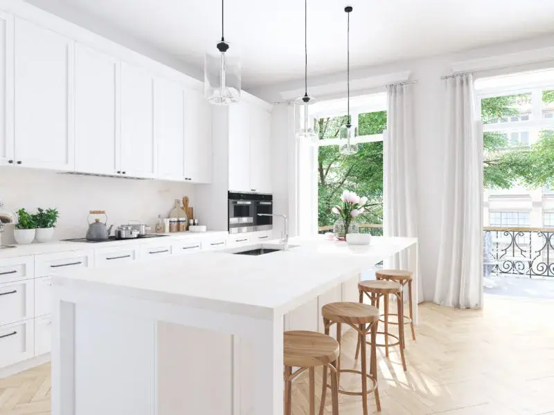 An image of a white kitchen with shaker cabinets, wooden bar stools, and floor to ceiling windows with grey curtains.